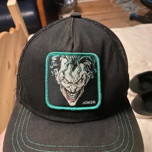Svart keps med Joker-motiv - Snygg svart keps med broderad patch av Jokern framtill. Kepsen har gröna kontrastsömmar och mesh på sidorna för extra ventilation. Perfekt för dig som gillar serietidningsstil och vill sticka ut med en unik accessoar.