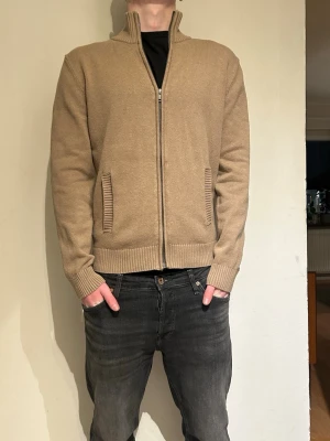 Beige stickad ziptröja - Snygg beige stickad ziptröja med hög krage och ribbade muddar. Tröjan har två fickor framtill och dragkedja hela vägen upp. Tröjan har inga defekter. Skick 8/10.