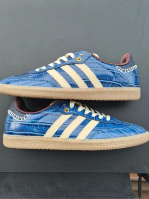 Wales Bonner sambas  - Tja! Säljer dessaUnika blå Wales bonners från Adidas med krokodil-liknande mönster, klassiska vita stripes på sidorna och beige sula. Skorna har vita skosnören, gulddetaljer vid snörningen och brun insida. Perfekt för dig som vill sticka ut med din stil.