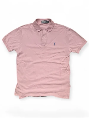 Rosa pikétröja från Polo Ralph Lauren - Klassisk rosa pikétröja från Polo Ralph Lauren med korta ärmar och krage. Tröjan har knappar vid halsen och den ikoniska broderade loggan i blått på bröstet. Perfekt till jeans eller shorts för en avslappnad men snygg look.