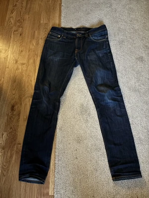 Mörkblå jeans från Nudie Jeans - Säljer ett par mörkblå jeans från Nudie Jeans med klassisk femficksmodell och kontrastsömmar. Jeansen har rak passform och är tillverkade i denim med snygg tvätt. Perfekta för dig som gillar stilrena och tidlösa jeans. Storlek W31 L32