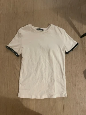 Vit t-shirt från zara - Vit t-shirt från Zara med korta ärmar och snygga pärldetaljer i svart och vitt vid ärmsluten. Enkel och stilren modell i mjuk bomull, perfekt för en trendig vardagslook. 