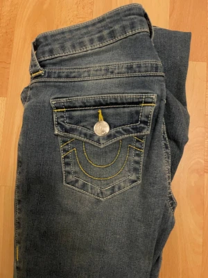 True Religion blå bootcut jeans - Säljer ett par blå jeans från True Religion, modell BECCA Mid Rise Bootcut. Jeansen har snygga kontrastsömmar i gult, dekorativ ficka bak och silverfärgade knappar. Klassisk bootcut passform och tillverkade i denim. Perfekta för en cool och avslappnad stil.