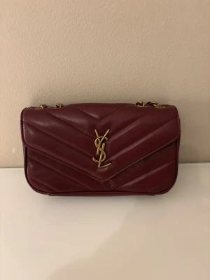 Vinröd YSL handväska i läder - Säljer en vinröd handväska från Yves Saint Laurent i quiltat läder med guldiga detaljer och kedjerem. Väskan har klassisk YSL-logga framtill och en stilren, rektangulär form. Perfekt för dig som vill ha en lyxig accessoar med ikonisk design.
