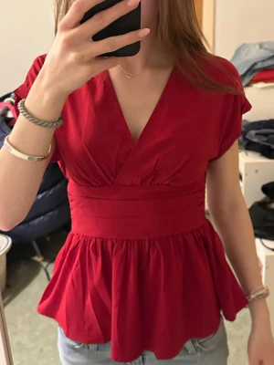 Röd omlottopp med v-ringning - Snygg röd topp med omlott-design och v-ringning. Modellen har korta ärmar, markerad midja och volangig nederdel som ger en feminin siluett. 