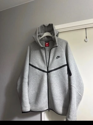 Grå Nike Tech Fleece hoodie - Snygg grå Nike Tech Fleece hoodie med svart dragkedja och svarta detaljer. Har huva, fickor på sidorna och en extra ficka med dragkedja på ärmen. Nike-logga broderad på bröstet. Perfekt för en sportig och stilren look.