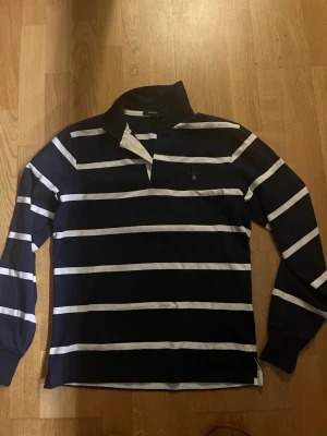 Randig långärmad skjorta från Gant - Mörkblå långärmad skjorta från Gant med vita horisontella ränder. Klassisk krage och broderad logga på bröstet. Perfekt för en avslappnad men stilren look.