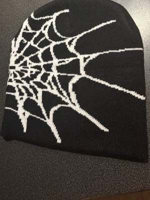 Svart mössa med spindelnätsmönster - Märket Spider Cool svart mössa med ett stort vitt spindelnätsmönster på sidan. Mössan är stickad och har en tight passform som sitter snyggt på huvudet. Perfekt för dig som gillar alternativ stil eller vill sticka ut lite extra. Materialet känns mjukt och bekvämt.