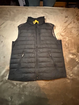 Svart dunväst från Parajumpers - Snygg svart dunväst från Parajumpers med gul detalj vid kragen och två dragkedjeförsedda fickor framtill. Västen har hög krage och quiltad design som ger en sportig look. Perfekt för lager-på-lager och håller dig varm under kyliga dagar.