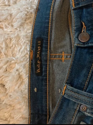 Mörkblå jeans från Nudie Jeans - Säljer ett par mörkblå jeans från Nudie Jeans med klassisk femficksmodell och raka ben. Jeansen har kontrastsömmar och signaturbroderi på bakfickorna. Tillverkade i slitstark denim, perfekta för en avslappnad och snygg stil.