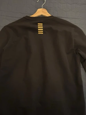 Svart sweatshirt från EA7 Emporio Armani - Snygg svart sweatshirt från EA7 Emporio Armani med gul logga på bröstet och gulrandig detalj i nacken. Tröjan har rund halsringning och långa ärmar. Perfekt för en stilren och sportig look.