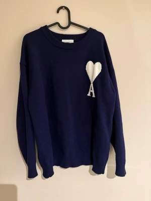 Mörkblå stickad tröja Ami Paris - Mörkblå stickad tröja från Ami Paris med ikoniskt vitt hjärta och bokstaven A broderat på bröstet. Tröjan har rund halsringning, långa ärmar och ribbade muddar. Perfekt för dig som gillar stilrena detaljer och klassisk design.