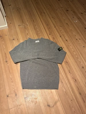 Grå stickad tröja från Stone Island - Säljer en stilren grå stickad tröja från Stone Island med klassisk rund hals och ribbade muddar. Tröjan har den ikoniska Stone Island-loggan på vänster ärm. Perfekt för dig som gillar streetwear och enkel design.
