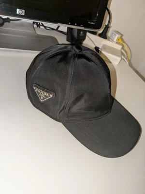 Svart keps från Prada i nylon - Snygg svart keps från Prada i glansigt nylonmaterial med klassisk böjd skärm och justerbar rem bak. På sidan sitter Pradas ikoniska metallogo i trekant. Perfekt accessoar för en trendig och stilren look.