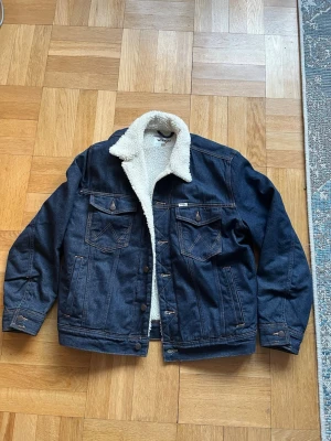 Wrangler denim jacket L - Säljer en mörkblå jeansjacka från Wrangler med vitt teddyfoder. Klassisk modell med två bröstfickor, knappstängning och kontrastsömmar. Perfekt för dig som vill ha en tidlös och snygg jacka med extra värme och cool vintagekänsla. Nypris 1199kr men säljer då den är för stor för mig