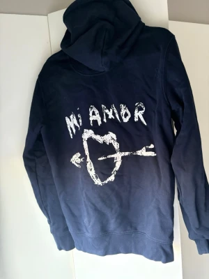 Marinblå hoodie med tryck Mi Amor - Snygg marinblå hoodie med stor ficka framtill och huva med dragsko. På ryggen finns ett vitt tryck med texten 'Mi Amor' och ett hjärta genomborrat av en pil. Perfekt för dig som gillar streetwear och vill sticka ut lite extra.