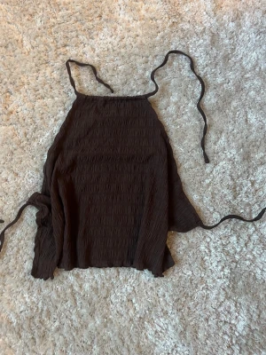Brun ärmlös linnetopp med knyt - Säljer en brun ärmlös linnetopp med smockad struktur och tunna knytband i halsen och ryggen. Perfekt för dig som gillar en enkel men trendig look. Toppen har en loose passform och är superenkel att styla till olika outfits. Öppen i ryggen 