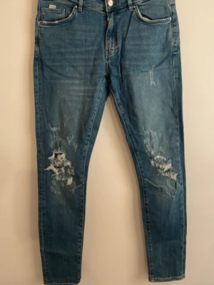 Jeans från Karve - Säljer ett par blå jeans med skinny fit och slitningar samt hål på båda knäna. Klassisk femficksmodell med dragkedja och knapp. Jeansen har en cool, avslappnad look och är perfekta för dig som gillar streetstyle.