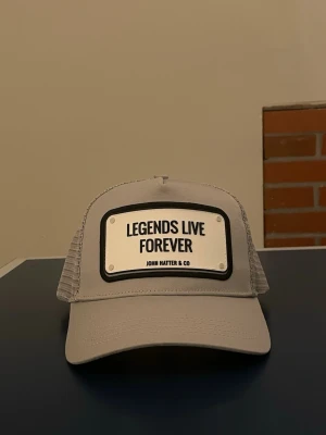 Grå keps John Hatter & Co - Snygg grå keps från John Hatter & Co med texten 'Legends Live Forever' framtill. Kepsen har klassisk böjd skärm och mesh på sidorna och bak för extra ventilation. Perfekt accessoar för en cool och avslappnad stil.