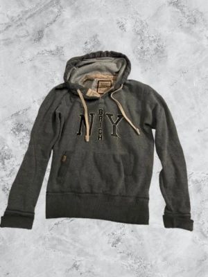 Grå hoodie från Better Rich NY - Mysig grå hoodie från Better Rich med broderad text 'NY BRICH' på bröstet. Hoodien har beige dragsko, känguruficka och ribbade muddar. Insidan är mjukt fodrad för extra komfort. Perfekt för en avslappnad streetstyle-look.