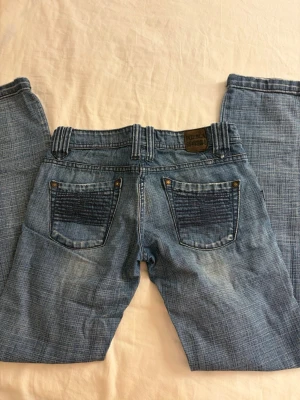 Blå jeans från Pueblo med mönstrade fickor - Snygga blå lågmidjade jeans från Pueblo med unikt ”rutmönster” i tyget och coola, mönstrade fickor både fram och bak. Jeansen har raka ben, bälteshällor och klassisk knappgylf. Perfekta för dig som vill sticka ut lite extra med din denimstil. Måtten finns på bilderna och priset är diskuterbart💕💕