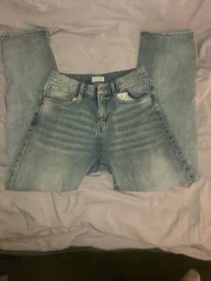 Ljusblå jeans från Lindex - Säljer ett par ljusblå jeans från Lindex med klassisk femficksdesign och raka ben. Jeansen har en snygg tvättad look med slitningar och tydliga whiskers vid låren. Perfekta för en avslappnad och trendig stil.