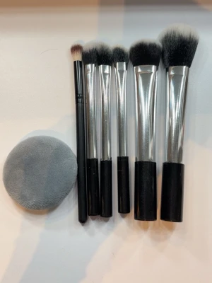 Set med sminkborstar och puff - Säljer ett set med fem sminkborstar och en mjuk, rund puderpuff. Borstarna har svarta handtag, silverfärgade detaljer och täta, mjuka borst i svart och vitt. Perfekt för applicering av foundation, puder och ögonskugga. Puffen är grå och superlen.