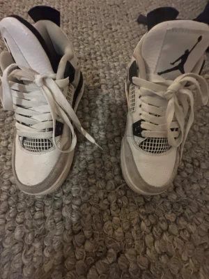 Vita Air Jordan 4 Retro sneakers - Snygga Air Jordan 4 Retro sneakers i vitt med svarta detaljer och klassisk Jumpman-logga på plösen och sulan. Skorna har snörning och meshpaneler på ovandelen. Perfekta för dig som gillar streetwear och ikoniska sneakers.