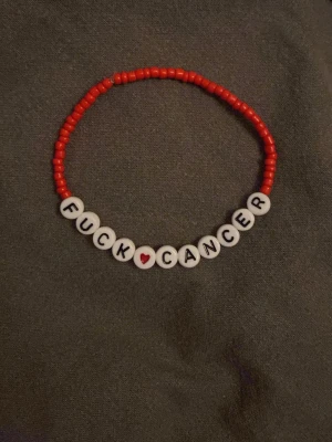Rött pärlarmband med text - Handgjort armband med röda små pärlor och vita bokstavspärlor som stavar 'FUCK ❤️ CANCER'. Hjärtat är rött och ger en extra detalj. Perfekt för dig som vill bära ett statement och visa stöd.