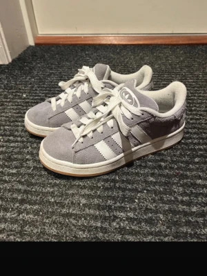 Grå Adidas sneakers med vita ränder - Snygga grå sneakers från Adidas med klassiska vita ränder på sidorna och vit sula. Skorna har en låg profil och är gjorda i mockaliknande material med vita skosnören och Adidas-logga på plösen. Perfekta för en stilren och avslappnad look.