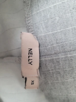 Grå kostymbyxa från Nelly - Säljer ett par ljusgrå kostymbyxor från nelly med diskreta vertikala ränder. De har en mjuk och stretchig passform med bred resår i midjan. Perfekta för en avslappnad och trendig look.