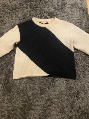 Beige och svart stickad tröja - !!Kommer att tvättas!!Stickad tröja med rund hals i beige och svart, där färgerna är diagonalt delade över bröstet. Långärmad och mjuk, perfekt för kalla dagar. Ingen synlig logga eller märke på tröjan. Passar dig som gillar enkel och stilren design.
