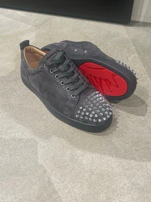 Gråa Louboutin sneakers med nitar - Snygga gråa sneakers i mocka från Christian Louboutin med ikonisk röd sula och silvriga nitar på tån. Skorna har svarta snören och låg profil, perfekt för dig som vill sticka ut med en edgy stil. Klassisk Louboutin-design med lyxig känsla.