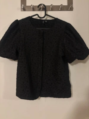 Svart puffärm blus med struktur - Snygg svart blus med puffiga korta ärmar och rund halsringning. Blusen har ett strukturerat tyg med diskret blommönster. Går att både klä upp till fest eller klä ner till skolan. Blusen är aldrig använd.