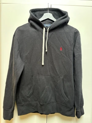 Svart hoodie från Polo Ralph Lauren - Klassisk svart hoodie från Polo Ralph Lauren med vit dragsko och röd broderad logga på bröstet. Hoodien har känguruficka framtill och mjukt bomullsmaterial som känns skönt mot huden. Perfekt för en avslappnad och stilren look.