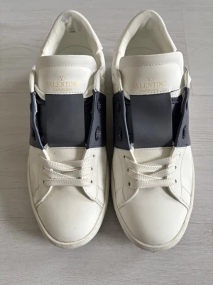 Varltino skor - Snygga vita sneakers från Valentino Garavani med svart detalj över sidan och klassiska nitar på hälen. Skorna är låga och har vita snören samt en chunky sula. Tillverkade i läder för en exklusiv känsla och stilren look. OBS!!! Priset är rörligt