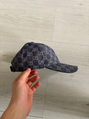 Blå Gucci Keps - Säljer en mörkblå keps från Gucci med klassiskt GG-monogram i grått över hela kepsen. Justerbar rem bak i matchande färg och gulddetalj. Snygg och stilren modell som passar till streetwear och trendiga outfits. OBS!!! Priset är rörligt