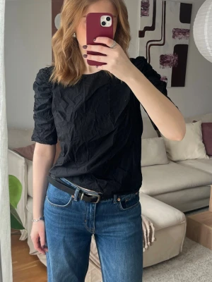 Svart topp med puffärm - Snygg svart topp med puffiga, rynkade ärmar och rund halsringning. Modellen är rak och materialet känns lätt och bekvämt. Perfekt att styla med jeans eller kjol för en trendig look.