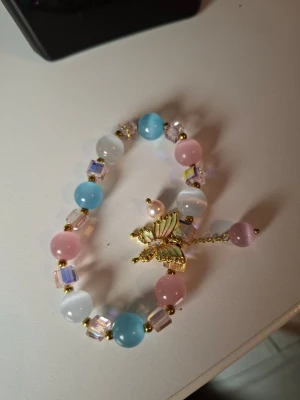 Färgglatt pärlarmband med fjäril - Sött armband med pärlor i pastellfärger som rosa, blått och vitt samt kubformade glaspärlor. Armbandet har en guldfärgad fjärilsberlock i mitten och små gulddetaljer mellan pärlorna. Perfekt accessoar för dig som gillar gulliga och färgglada smycken.