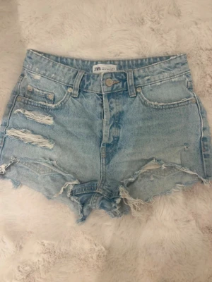 Ljusa blåa jeansshorts Zara - Säljer ett par ljusblåa jeansshorts från Zara med slitna detaljer och råa kanter. Shortsen har klassisk femficksdesign, knappgylf och en relaxed fit. 