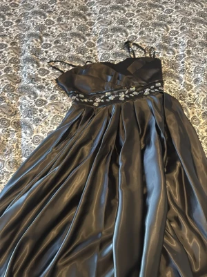 Svart långklänning med blommiga detaljer - Elegant svart långklänning med smala axelband och v-ringad överdel. Klänningen har ett glansigt satinliknande material och ett brett midjeband dekorerat med broderade blommor i silver och vitt. Perfekt för speciella tillfällen.