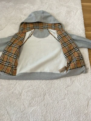 Grå hoodie med Burberry-rutor - Snygg grå hoodie med dragkedja från Burberry. Insidan av huvan och jackan har det klassiska Burberry-rutiga mönstret i beige, svart och rött. Mjuk bomullskänsla, två fickor fram och dragsko i huvan. Perfekt för en stilren och trendig look.