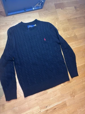 Svart kabelstickad tröja från Polo Ralph Lauren - Kabelstickad svart tröja från Polo Ralph Lauren med rund hals och lång ärm. Tröjan har ett klassiskt rött broderat logotyp på bröstet och ribbade muddar vid ärmslut och nederkant. Perfekt för dig som gillar stilrena och tidlösa plagg. Aldrig använd skriv vid frågor och funderingar 