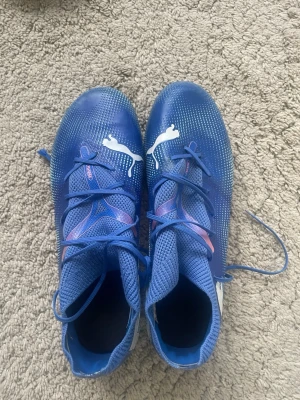 Blåa Puma Ultra fotbollsskor - Säljer ett par blåa Puma Ultra fotbollsskor med vita detaljer och logga. Skorna har snörning, ovandel i syntetmaterial och en ljusgrön sula med fasta dobbar för bra grepp på planen. Perfekta för dig som vill ha lätta och snabba skor på matchen. Storlek 42,5. Säljer pga. att de är för små för mig. 