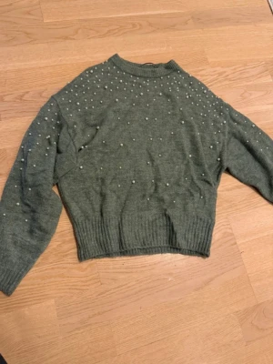 Grön stickad tröja med pärlor - Mysig grön stickad tröja med pärldetaljer över axlar och bröst. Tröjan har en rak, något kortare passform, ribbade muddar och rund halsringning. Perfekt för dig som vill ha något extra till din vardagsstil.