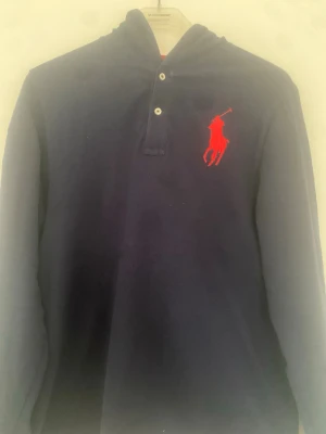 Marinblå hoodie från Polo Ralph Lauren - Snygg marinblå hoodie från Polo Ralph Lauren med stor röd logga på bröstet och siffran 3 i rött på ärmen. Hoodien har knappar vid halsen och en huva med röd insida. Perfekt för en sportig och avslappnad stil.