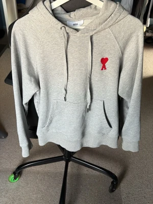 Grå hoodie från Ami - Snygg grå hoodie fråån Ami. På bröstet finns ett broderat rött hjärta med A. Perfekt för en chill och stilren look. Mjuk bomullsblandning och avslappnad passform. Lite fläckig på ryggen (se bild 2) därför det låga priset