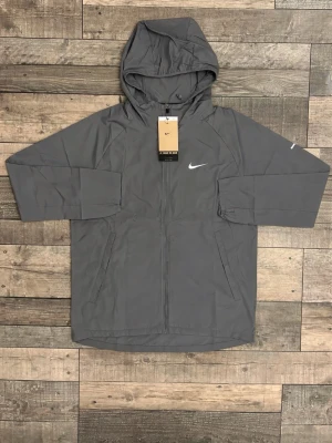 Grå vindjacka från Nike med huva - Snygg grå vindjacka från Nike med dragkedja framtill, justerbar huva och två sidofickor. Jackan har Nike-logga på bröstet och är tillverkad i ett lätt, vindtätt material. Perfekt för blåsiga dagar och sportiga outfits.