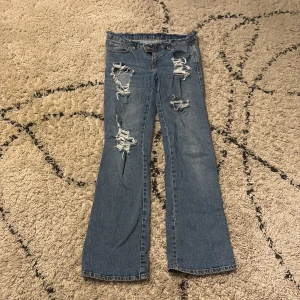 Blå slitna bootcut jeans med spets - Säljer ett par blå bootcut jeans med slitningar och hål framtill samt vit spetsdetalj på bakfickan. Jeansen har väldigt låg midja och rå kant upptill för en extra edgy look. Perfekta för dig som gillar en trendig och cool stil! Ny köpta från lager157 men har sedan gjort hål, klippt av kanten och sytt på spets. Passar mig perfekt i längden som är 173!