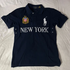 Mörkblå pikétröja Polo Ralph Lauren - Mörkblå pikétröja från Polo Ralph Lauren med broderad logga, emblem och texten 'NEW YORK' framtill. Klassisk krage, korta ärmar och knappar vid axlarna. Perfekt för en sportig och stilren look.